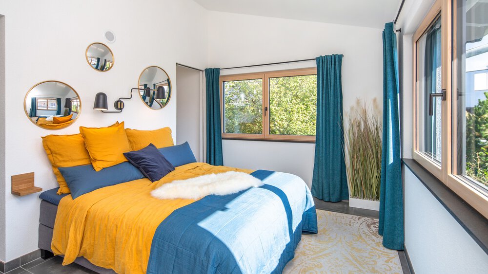 Helles Schlafzimmer mit gelber und blauer Bettwäsche, runden Spiegeln an der Wand und einem großen Fenster mit blaugrünen Vorhängen.