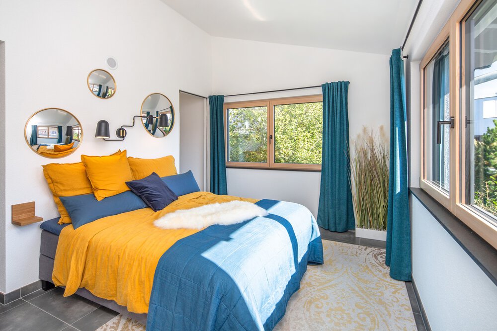 Helles Schlafzimmer mit gelber und blauer Bettwäsche, runden Spiegeln an der Wand und einem großen Fenster mit blaugrünen Vorhängen.