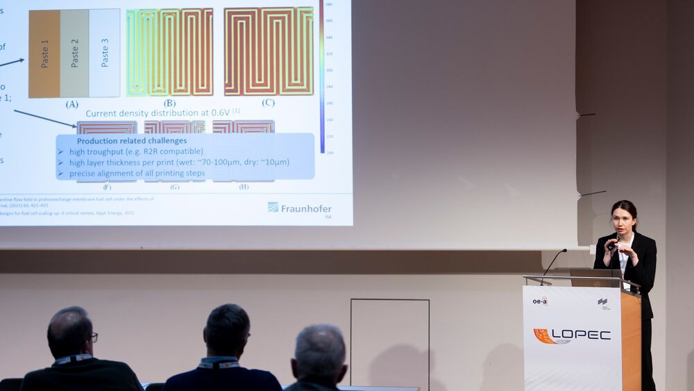 A woman stands on a stage at a lectern and gives a presentation to an audience with the help of a PowerPoint presentation.