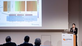A woman stands on a stage at a lectern and gives a presentation to an audience with the help of a PowerPoint presentation.