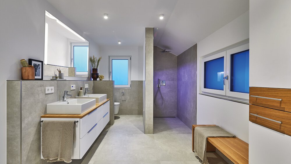Modernes Badezimmer mit Doppelwaschbecken, großer Dusche, Sitzbank und blau getönten Fenstern, beleuchtet durch Einbauleuchten.