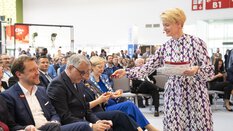 Eine Frau in einem gemusterten Kleid steht vor einem sitzenden Publikum und reicht einem der Anwesenden einen gefalteten roten Zettel.