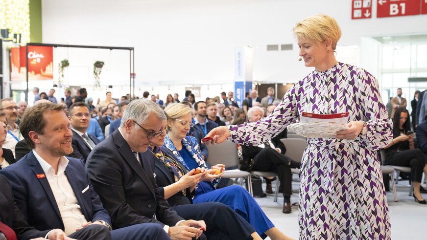 Eine Frau in einem gemusterten Kleid steht vor einem sitzenden Publikum und reicht einem der Anwesenden einen gefalteten roten Zettel.