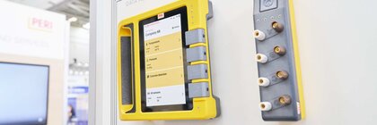 A yellow, industrial tablet for data acquisition is displayed on a white wall at a trade fair stand.