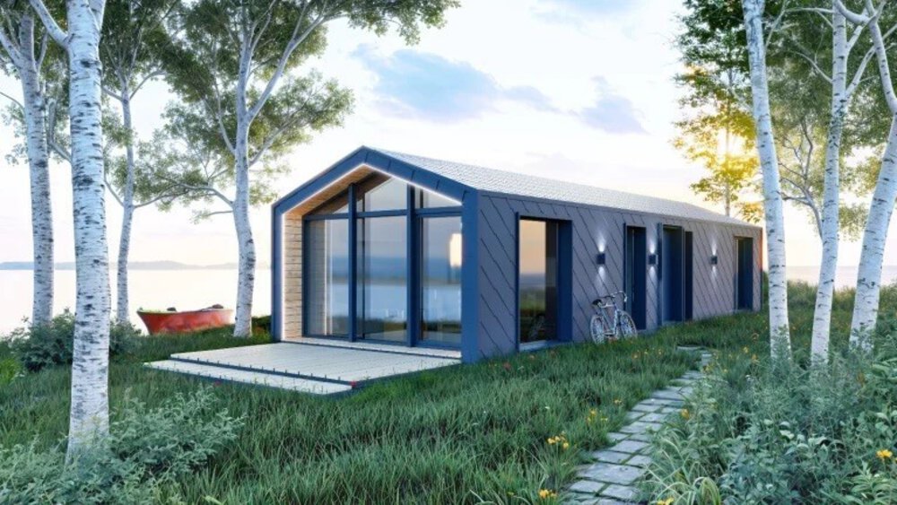 Ein längliches Tinyhouse mit großen Fensterfronten und einem Fahrrad stehen zwischen Birken an einem See unter einem bewölkten Himmel.