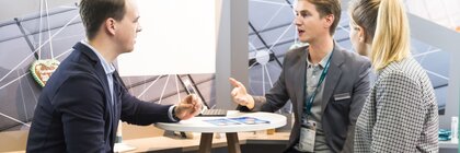 Three people in business attire are talking at a small round table with flyers and a laptop.