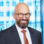 Ein lächelnder, kahlköpfiger Mann mit Brille und Bart in Anzug und Krawatte.