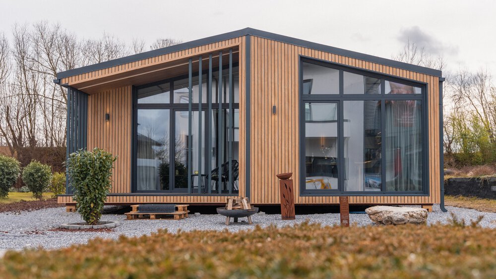 Außenfassade eines hölzernen Bungalows mit großen Glasfronten, Außenbeleuchtung und einem Steingarten mit einer Feuerstelle.