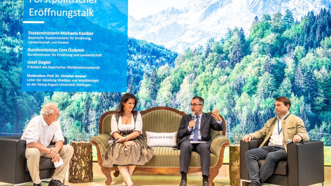 Three men and a woman sit on a stage and discuss, on the left a blue banner with event details for the 