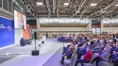 Links befindet sich eine Bühne in einer großen Messehalle, auf der zwei Personen bei einem Vortrag sprechen. Rechts sitzt die Publikumsmenge.