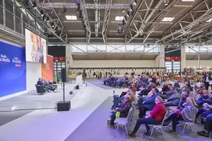 Links befindet sich eine Bühne in einer großen Messehalle, auf der zwei Personen bei einem Vortrag sprechen. Rechts sitzt die Publikumsmenge.