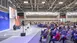 On the left is a stage in a large exhibition hall where two people are giving a talk. On the right is the audience.