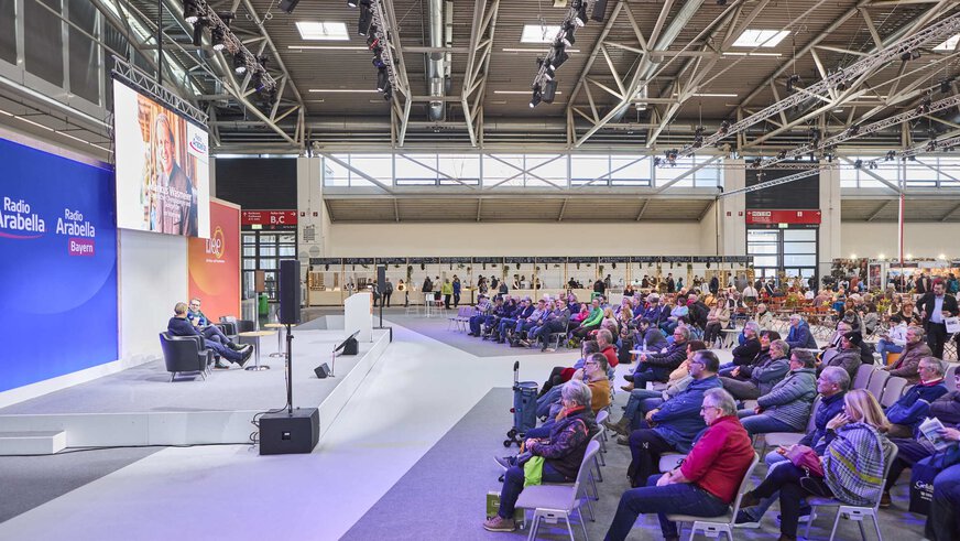 Links befindet sich eine Bühne in einer großen Messehalle, auf der zwei Personen bei einem Vortrag sprechen. Rechts sitzt die Publikumsmenge.