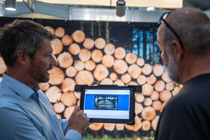 Two men are looking at a tablet in front of a stack of logs