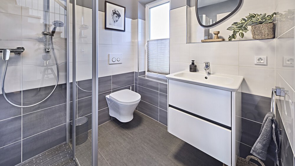 Dieses moderne Gästebad im Musterhaus von KAMPA verfügt über eine elegante Glasdusche, eine wandmontierte Toilette und ein Waschbecken mit Spiegel, alles mit dekorativen Pflanzen verziert.