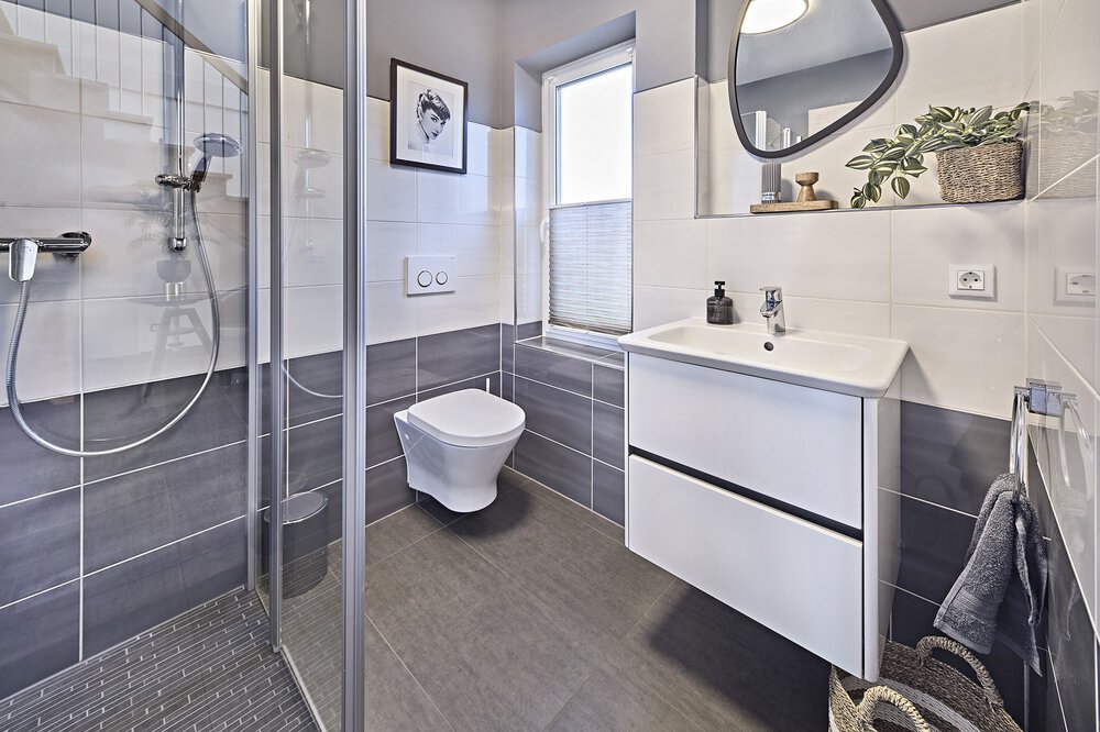 Dieses moderne Gästebad im Musterhaus von KAMPA verfügt über eine elegante Glasdusche, eine wandmontierte Toilette und ein Waschbecken mit Spiegel, alles mit dekorativen Pflanzen verziert.