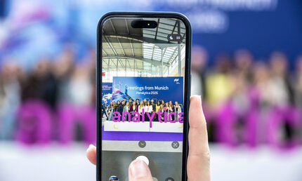 Hand mit Smartphone - Gruppenfoto vor analytica Schriftzug