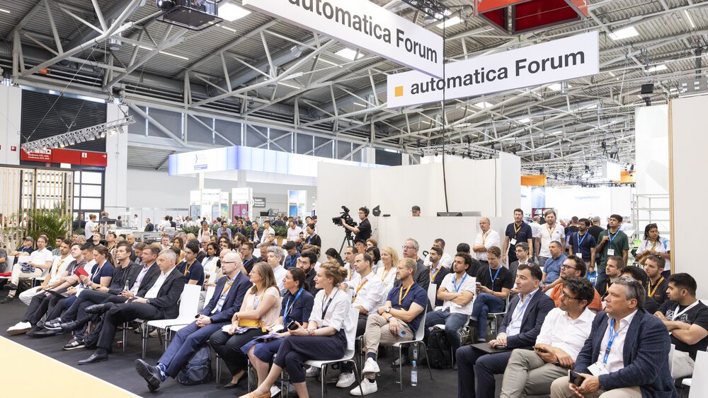 Ein interessiertes Publikum lauscht einem Vortrag im automatica Forum. Im Hintergrund ist eine gut besuchte Messehalle zu sehen.