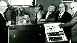 Schwarz-weißes Foto von einer früheren productronica. Mehrere Männer in Anzügen stehen um eine Maschine herum, drücken Knöpfe und schauen interessiert.