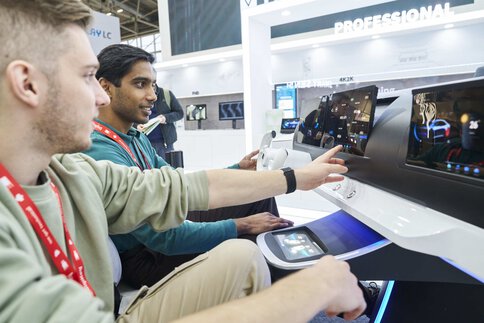 Two people sit in front of a futuristic dashboard exhibited at a trade fair stand and interact with the technology.