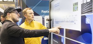 Two men discuss information on a large touchscreen display at a trade fair stand.