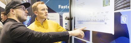 Two men discuss information on a large touchscreen display at a trade fair stand.