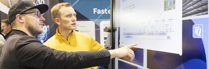 Two men discuss information on a large touchscreen display at a trade fair stand.