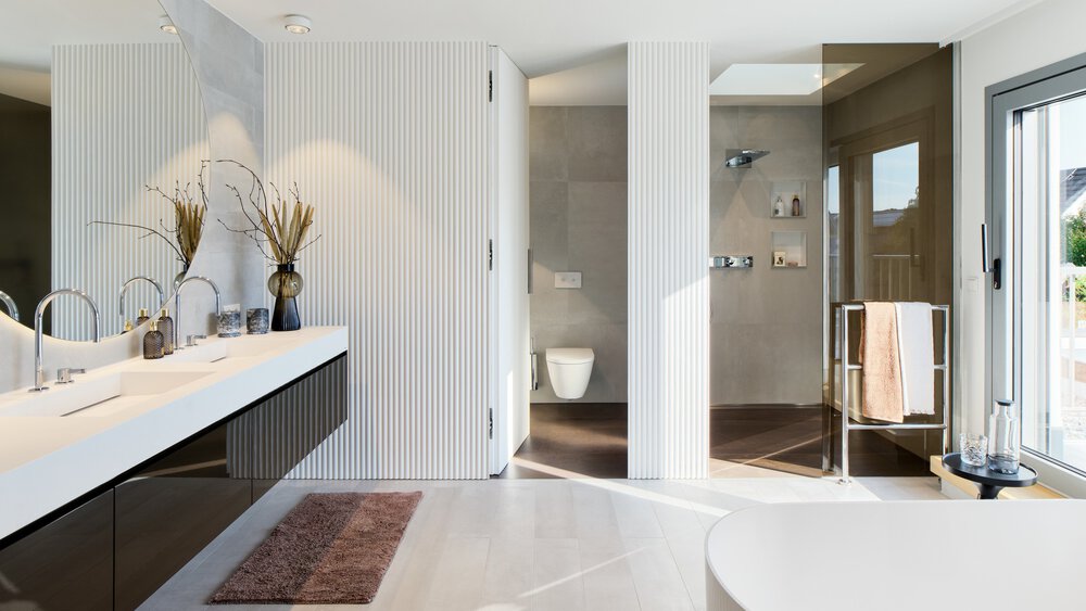 Modernes Badezimmer mit weißer Waschtischplatte, Vasendekor, zwei Waschbecken, einer Dusche und einer Toilette. Großes Fenster und helle Beleuchtung.