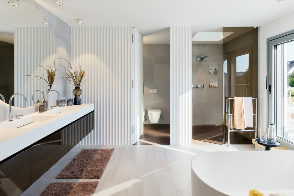 Modernes Badezimmer mit weißer Waschtischplatte, Vasendekor, zwei Waschbecken, einer Dusche und einer Toilette. Großes Fenster und helle Beleuchtung.