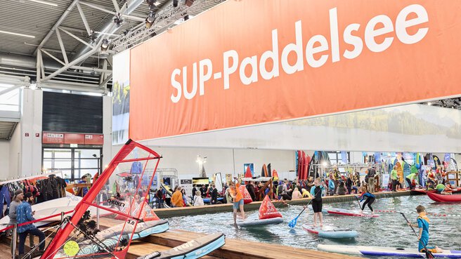 Indoor water pool at the f.re.e trade fair with children and adults stand-up paddling under a large “SUP paddle lake” banner.