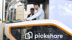 Ein Mann sitzt im Führerhaus eines kleinen Transportfahrzeugs mit Fahrradlenker. Ein anderer Besucher schaut ihm lächelnd über die Schulter.
