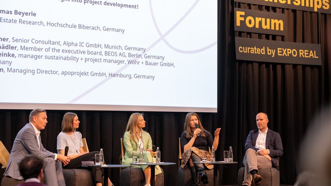 Five people are sitting on a panel discussion stage, behind them is a screen and a sign saying 
