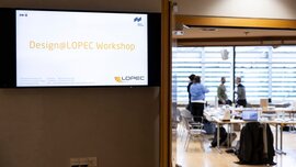 Screen displaying Design@LOPEC Workshop sign with view into a workshop room and participants
