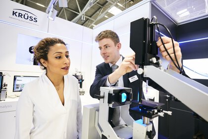 Ein Unternehmensmann zeigt einer Unternehmensfrau ein Gerät iaus dme Bereich der Grenzflächenanalytik am Messestand