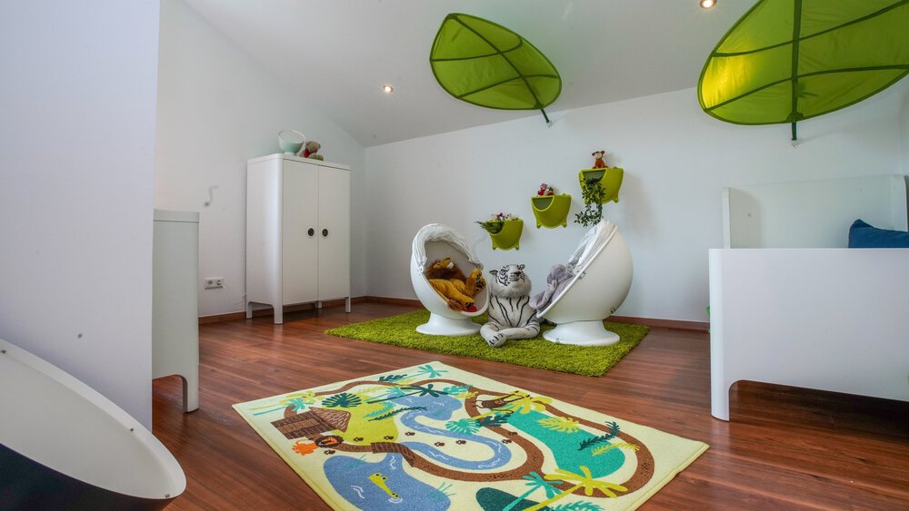 Verspielters Kinderzimmer mit grünem Blätterdekor an der Decke, verspielten Stühlen, weißen Möbeln und einem farbenfrohen Teppich mit Dschungelmotiv.