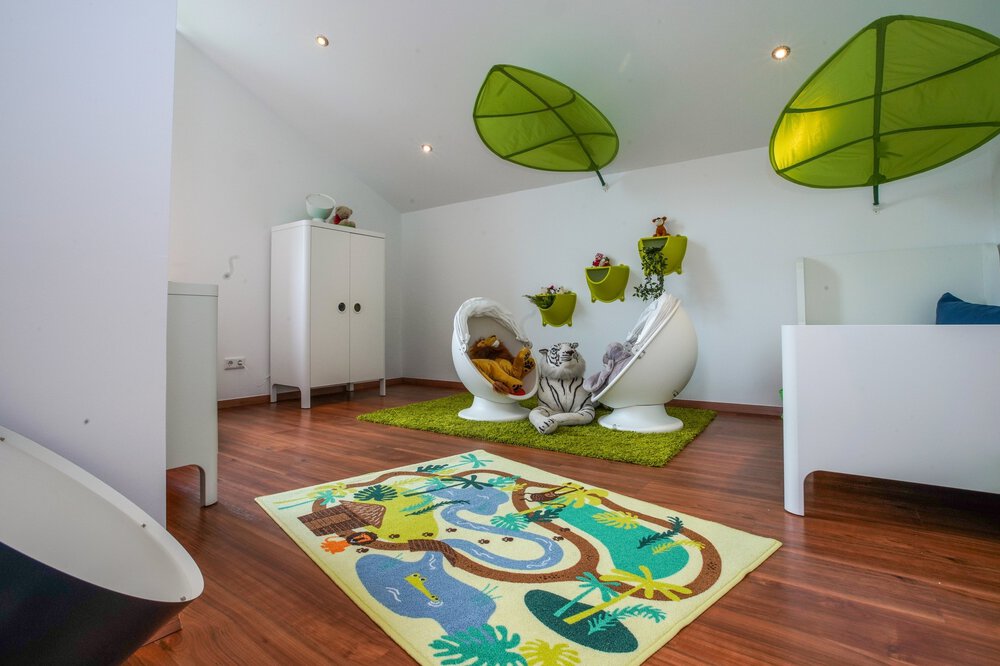 Verspielters Kinderzimmer mit grünem Blätterdekor an der Decke, verspielten Stühlen, weißen Möbeln und einem farbenfrohen Teppich mit Dschungelmotiv.