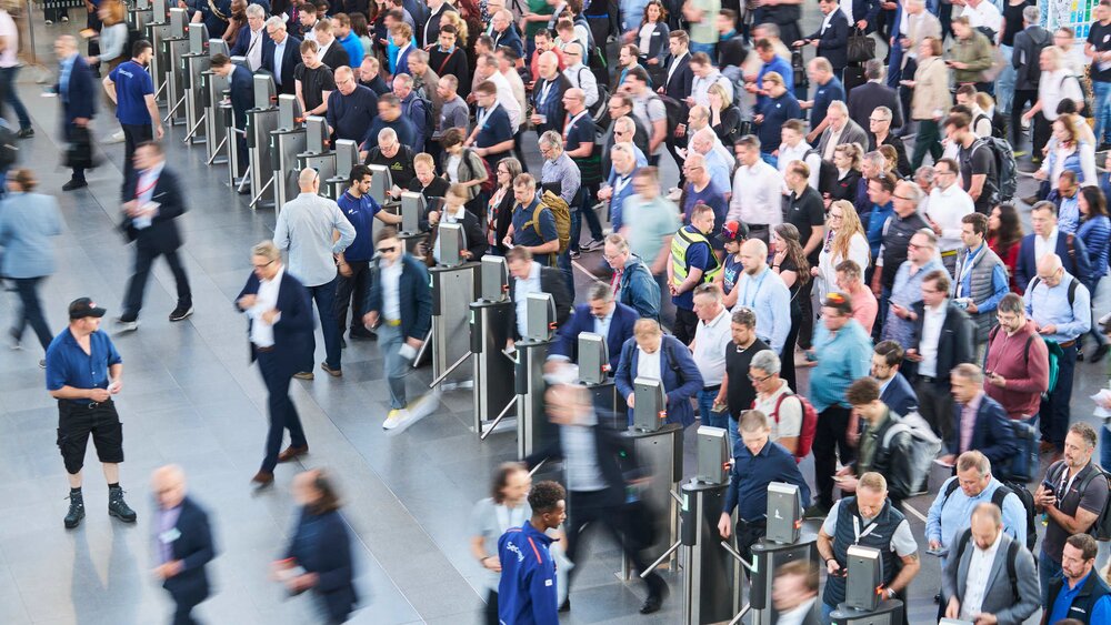 Overhead view of a dense crowd of trade fair visitors quickly passing through entrance controls and turnstiles.