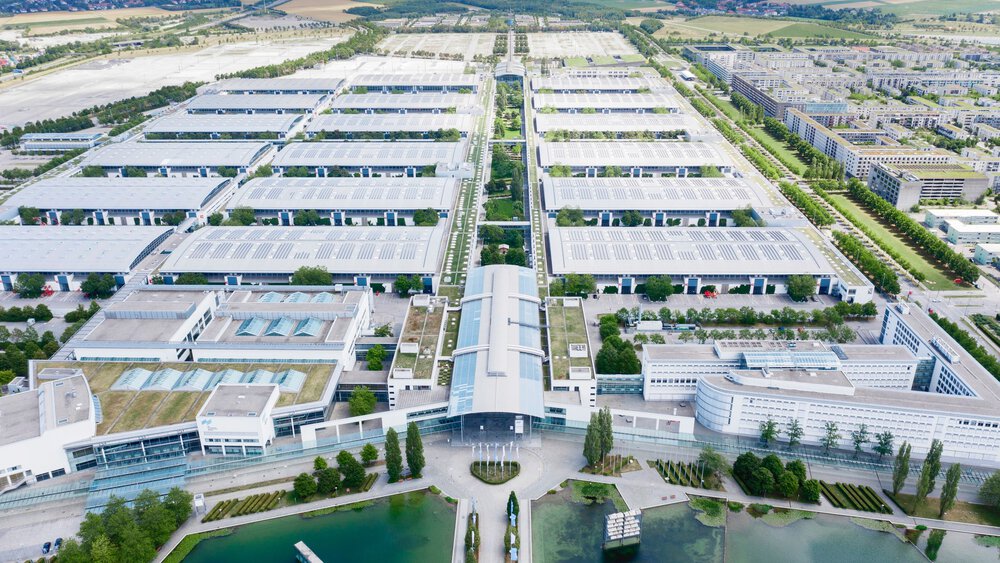 Ein Blick aus der Vogelperspektive über das gesamte Gelände der Messe München mit dem Messesee im Vordergrund, den zahlreichen Hallen und Feldern im Hintergrund.