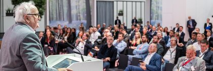 A speaker gives a presentation at a podium to an audience seated in an exhibition hall.