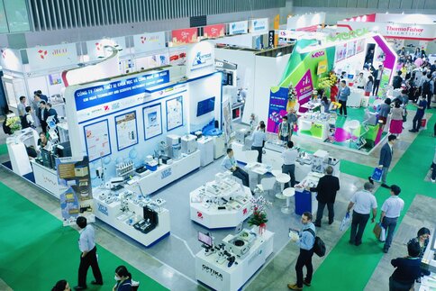 Top view of a well-attended trade fair with several colorful booths for laboratory and measurement technology, where attendees examine equipment and engage in consultation talks.