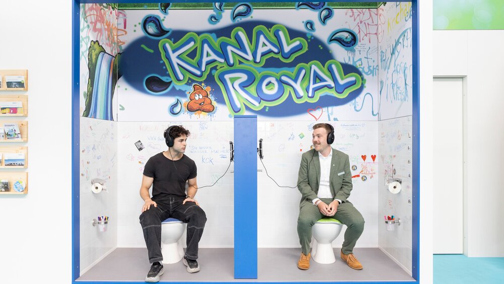 Two men wearing headphones sitting on mock-up toilets at a creatively designed trade fair booth labeled 