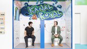 Zwei Männer sitzen mit Kopfhörern auf Toilettenattrappen in einem kreativ gestalteten Messestand mit der Aufschrift „Kanal Royal“.