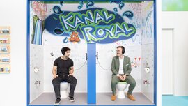 Two men wearing headphones sitting on mock-up toilets at a creatively designed trade fair booth labeled 