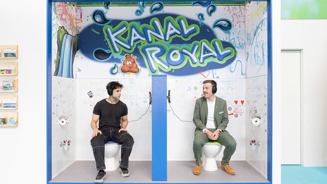Zwei Männer sitzen mit Kopfhörern auf Toilettenattrappen in einem kreativ gestalteten Messestand mit der Aufschrift „Kanal Royal“.
