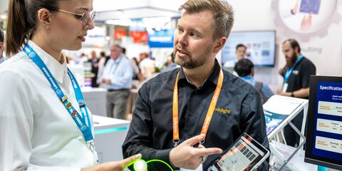 Two people are discussing technology at a trade fair. One is holding a device and the other is pointing at a tablet.