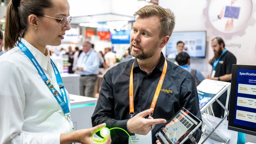 Zwei Personen diskutieren auf einer Messe über Technologie. Eine hält ein Gerät und die andere zeigt auf ein Tablet.