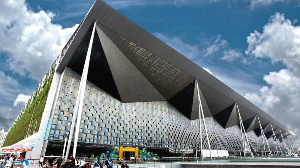 A large, modern building with angular architecture under a partly cloudy sky, in front of which people have gathered.