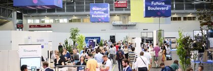Messehalle mit vielen Besucherinnen und Besuchern an Ständen und Sitzbereichen, über ihnen hängen große Displays mit der Aufschrift „Quantum Future Boulevard“.