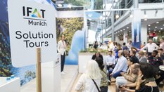 Hochgehaltenes Schild der IFAT Munich Solution Tours, dahinter sind unscharf einige Menschen zu sehen die einem Vortrag an einem Messestand lauschen.