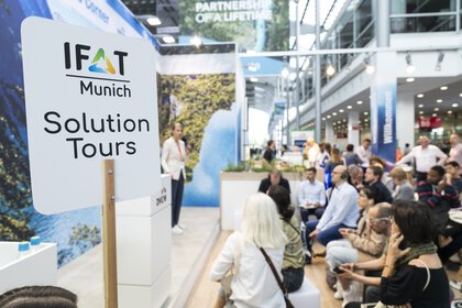 IFAT Munich Solution Tours sign held up high, behind it a few people can be seen out of focus listening to a presentation at an exhibition stand.